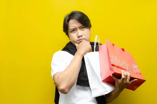 Alışveriş torbaları tutan düşünceli genç Asyalı adam, alışverişlerini düşünüyor. Alışveriş, karar verme ya da tüketici davranışı kavramları için ideal