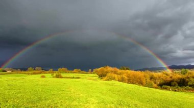 Bereketli yeşil tarlaların ve sonbahar renklerinde ağaçların üzerinde fırtınalı bir gökyüzünde nefes kesici bir gökkuşağı beliriyor. Sakin ama dramatik, seyahat ve tatil videolarında görkemli gösteriş için mükemmel.
