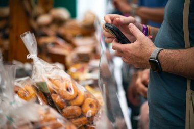 Bir adam akıllı telefonunu dikkatle kontrol ederken cazip çöreklerle dolu bir fırının önünde duruyor. Görüntü modern teknolojinin ve günlük yaşamın karışımını yakalıyor.