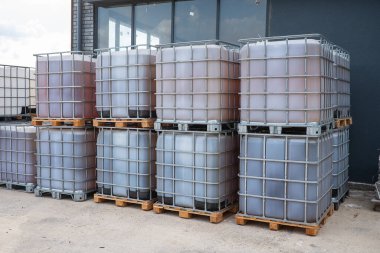 Açık hava sanayi ortamındaki ahşap paletlerin üzerine çeşitli renkli sıvılarla dolu büyük, dikdörtgen, metal çerçeveli kaplar yerleştirilmiştir.