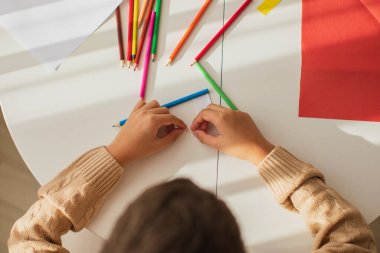 Child boy painting with colored pencils and making crafts and cardboard and colored paper uses glue and scissors