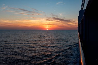 Bir geminin güvertesinden çekilen açık deniz üzerinde nefes kesici bir günbatımı görüntüsü. Huzurlu ufuk ve sıcak gökyüzü renkleri huzurlu ve resim gibi bir an yaratır..