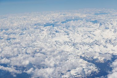 Uçsuz bucaksız kar örtülü dağların çarpıcı bir hava perspektifi, kabarık beyaz bulutlarla çevrili. Parlak mavi gökyüzü, bu yüksek irtifa manzarasının görkemli güzelliğini arttırır.