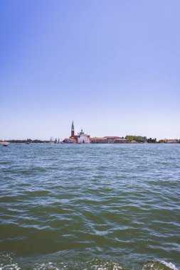 İtalya, Venedik 'teki San Giorgio Maggiore adasının sakin bir manzarası. İkonik kilise ve çan kulesi açık mavi gökyüzüne karşı yükseliyor. Venedik Gölünün sakin sularında ilerleyen bir motorlu tekne ön planda.