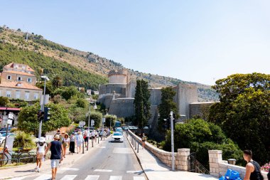 Yeşillik ve tepelerle çevrili tarihi bir kalenin yüksek ortaçağ duvarları yakınında canlı bir sokak manzarası. Turistler ve yerliler, zengin kültür mirasının tadını çıkararak yürüyüş yolunda yürüyorlar..