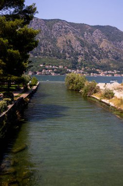 Karadağ 'ın Kotor kentinde yemyeşil bir alanla çevrili barışçıl bir kanal ve arka plandaki yüksek dağların çarpıcı manzarasını sunuyor. Huzurlu sular ve körfezin karşısındaki büyüleyici evler resim gibi ve rahatlatıcı bir manzara yaratıyor.