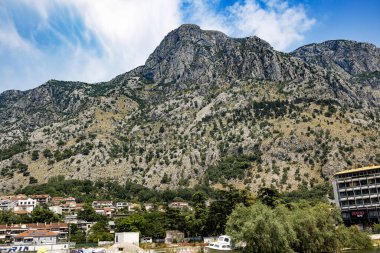 Karadağ 'ın Kotor kentinde yemyeşil bir alanla çevrili barışçıl bir kanal ve arka plandaki yüksek dağların çarpıcı manzarasını sunuyor. Huzurlu sular ve körfezin karşısındaki büyüleyici evler resim gibi ve rahatlatıcı bir manzara yaratıyor.