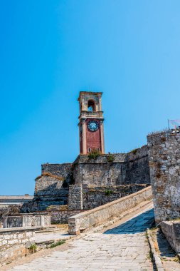 Yunanistan 'ın Corfu Adası' nın çarpıcı manzarası, yatlar ve yelkenlilerle dolu sakin bir körfezi gösteriyor. Ön plandaki gür yeşillik derin mavi deniz ve uzak tepelerle çok güzel tezat oluşturuyor.