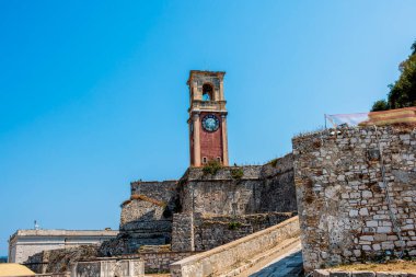 Yunanistan 'ın Corfu Adası' nın çarpıcı manzarası, yatlar ve yelkenlilerle dolu sakin bir körfezi gösteriyor. Ön plandaki gür yeşillik derin mavi deniz ve uzak tepelerle çok güzel tezat oluşturuyor.