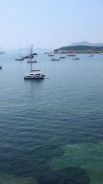 Yunanistan 'ın Corfu Adası' nın çarpıcı manzarası, yatlar ve yelkenlilerle dolu sakin bir körfezi gösteriyor. Ön plandaki gür yeşillik derin mavi deniz ve uzak tepelerle çok güzel tezat oluşturuyor.