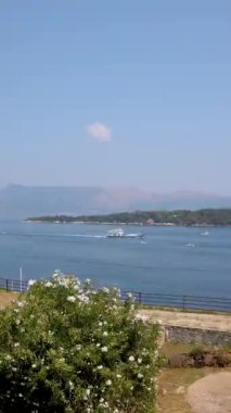 Yunanistan 'ın Corfu Adası' nın çarpıcı manzarası, yatlar ve yelkenlilerle dolu sakin bir körfezi gösteriyor. Ön plandaki gür yeşillik derin mavi deniz ve uzak tepelerle çok güzel tezat oluşturuyor.