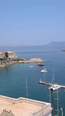 Yunanistan 'ın Corfu Adası' nın çarpıcı manzarası, yatlar ve yelkenlilerle dolu sakin bir körfezi gösteriyor. Ön plandaki gür yeşillik derin mavi deniz ve uzak tepelerle çok güzel tezat oluşturuyor.