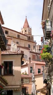 Maratea 'nın tarihi merkezinde renkli binalar, çiçekli balkonlar, dükkanlar ve arka planda kilise kulesi manzarası arasında turistlerin dolaştığı pitoresk bir ara sokak.