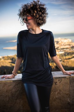 Kıvırcık saçlı, siyah tişörtlü arka planda bir şehirle gülümseyen genç bir kadın. Yüksek kalite fotoğraf