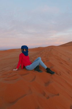 Gün batımında, Sahara 'da bir kum tepesinde berber eşarbıyla oturan genç bir kız. Yüksek kalite fotoğraf