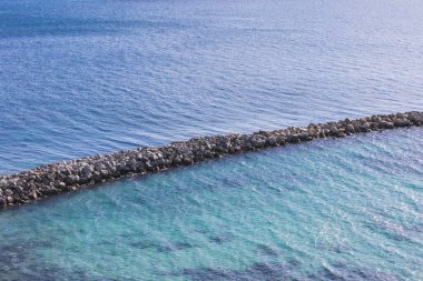 Akdeniz 'deki canlı turkuaz suları boyunca uzanan taştan bir dalgakıranın havadan görünüşü. Sakin sığ ve derin mavi okyanus arasındaki açık tezat sakin bir kıyı manzarası yaratır.. 