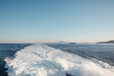Cebelitarık Boğazı 'nı geçen bir feribottan köpüklü bir iz. Ufukta kargo gemileri ve Afrika dağları görünüyor. Açık gökyüzü ve derin mavi deniz. Seyahat, ulaşım ve doğa konuları için mükemmel. Yüksek kalite fotoğraf