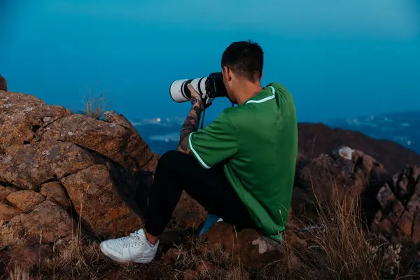 Kameralı ve teleskop mercekli bir adam kayalık dağdan alacakaranlıkta fotoğraf çekiyor. Macera, manzara ve arka planda manzaralı şehir ışıkları olan seyahat fotoğrafçılığı. Yüksek kalite fotoğraf