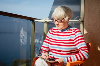 Deniz kenarında bir balkonda oturmuş dergilere bakıyor ve bulmaca çözüyor.