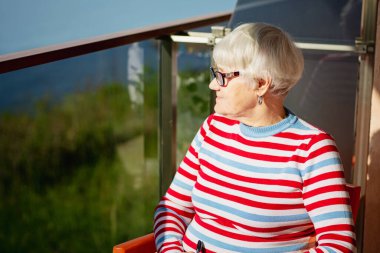 Elderly woman in glasses sitting on a balcony near the sea and looking at magazine, solving a crossword