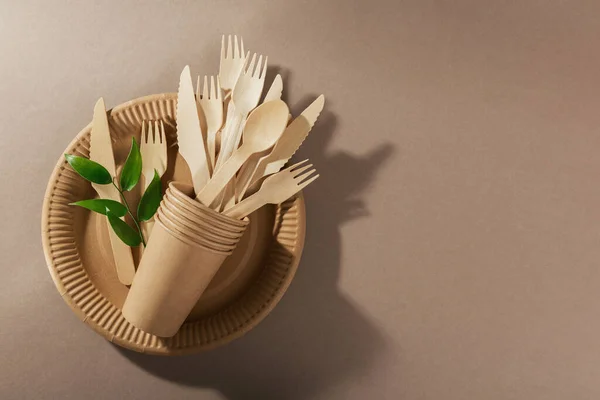 Eco-friendly disposable utensils made of bamboo wood and paper on a light beige background. Fork, knives, plates, paper cups and green leaves