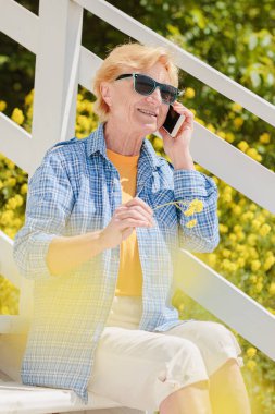 Mature attractive woman traveler sitting alone on the terrace of beach coffee shop in Bulgaria and using mobile phone. Active life of the elderly in retirement, active seniors