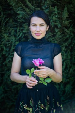 Young pretty woman in black clothes sits on a chair in the garden near a bush with a rose in her hands. Photography in a dark key