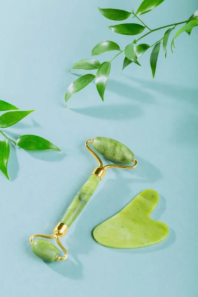 stock image Green jade face roller and gua sha scraper with leaves on light blue background. Massage tool for facial skin care, SPA self and beauty care treatment concept.