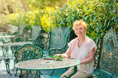 Mature attractive woman traveler sitting alone on the terrace of coffee shop in beautiful park in Bulgaria. Active life of the elderly in retirement, active seniors