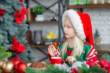 Tatlı bisküvi yiyen ve kupada sıcak kakao içen küçük tatlı bir kızın hayat tarzı portresi. Çocuk Noel süslemeli bir mutfakta oturuyor ve kırmızı Noel Baba şapkası takıyor.