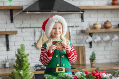 Noel Baba şapkalı genç kız. Mutfakta gülümsüyor ve Noel kurabiyesi yiyor.