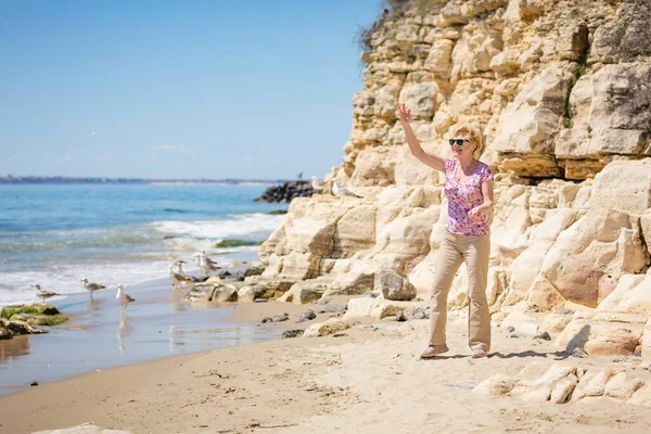 stock image Elderly woman in sunglasses feeds seagulls on the beach and laughs. Happy retirement life