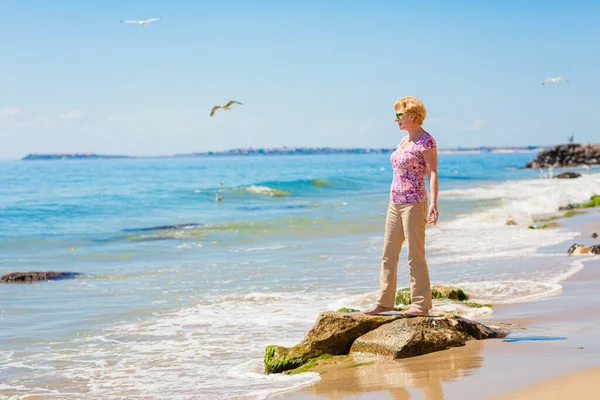 stock image Senior woman in sunglasses stands on the beach and looks at the sea. Old woman enjoying life near the sea