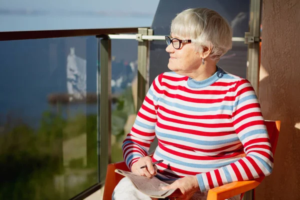 Elderly woman in glasses sitting on a balcony near the sea and looking at magazine, solving a crossword