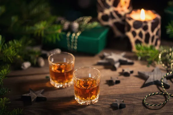 Two shot glasses of whiskey or bourbon with Holiday decoration on wooden background. New Year, Christmas and winter holidays whiskey mood concept