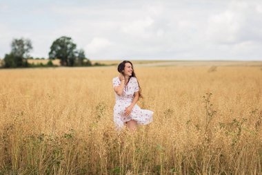 Young woman in white midi dress walking across the field with ripe golden ears of chickpeas. Beauty Romantic woman Outdoors