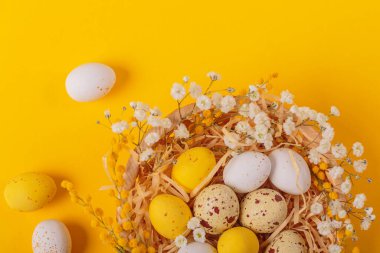 Easter candy chocolate eggs and almond sweets lying in a birds nest decorated with flowers and feathers on a yellow background. Happy Easter concept.