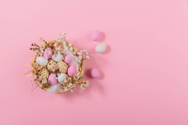 Easter candy chocolate eggs and almond sweets lying in a birds nest decorated with flowers and feathers on pink background. Happy Easter concept.