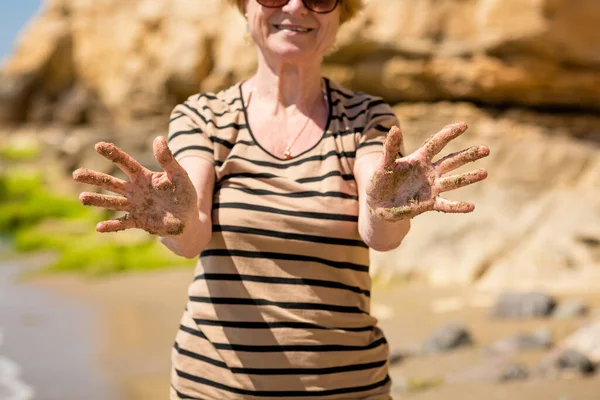 Elderly unrecognizable woman walking along the beach, showing her palm hands covered in sand, smiling and enjoying the moment