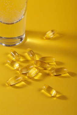 Diyet, beslenme, sağlıklı yeme konsepti. Sarı zemin üzerine sarı softgel kapsülleri dökmek için yağ dolu gıda takviyeleri ve bir bardak su..