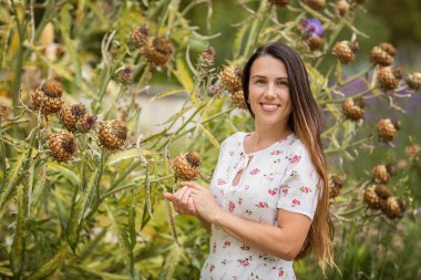 Young beautiful woman standing near big burdock plant and touching the leaves in old overgrown park