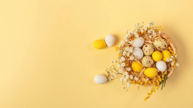 Easter candy chocolate eggs and almond sweets lying in a birds nest decorated with flowers and feathers on a yellow background. Top view long banner. Happy Easter concept.
