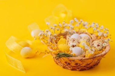 Easter candy chocolate eggs and almond sweets lying in a birds nest decorated with flowers and feathers on a yellow background. Happy Easter concept.