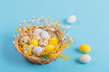 Easter candy chocolate eggs and almond sweets lying in a birds nest decorated with flowers and feathers on a blue background. Happy Easter concept.
