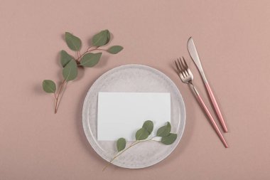 Blank white invitation stationery card on plate with eucalyptus leaves and cutlery on beige background. Flat lay, top view.