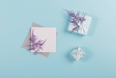Blank greeting card mockup with envelope, white blank paper, gift boxes and dried plant for birthday or holiday card with free space for message or design on blue background. Flat lay, top view.