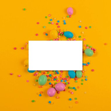 White blank paper on delicious chocolate colorful coated candy yellow background. Mock up card for congratulations or invitations. Top view, flat lay with free space for text