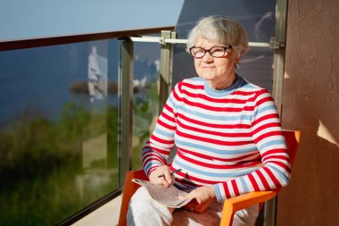 Senior woman in glasses sitting on a balcony near the sea and looking at magazine, solving a crossword