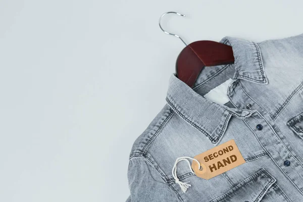 Gray denim shirt on hanger and tag with inscription second hand on white background. Second hand clothing shop. Circular fashion, eco friendly sustainable shopping, donation, thrift stores concept