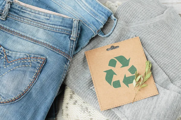 Sustainable still life with Blue denim jeans, gray pullover and craft paper card with Recycling symbol. Second hand apparel idea. Circular fashion, donation, charity concept. Top view